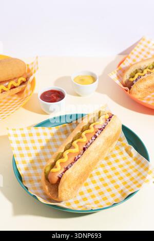 Ein klassischer Hot Dog, serviert in einem Brötchen mit einem Zickzack Senf, garniert mit Gurken und Zwiebeln. Perfekt für Fastfood-Liebhaber und Sommer-Picknicks, kombiniert Stockfoto