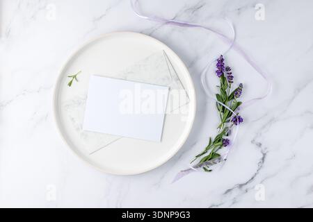 Mockup für Hochzeitseinladungskarten mit Blumenmuster. Leere Postkarte aus Papier, Vintage-Umschlag auf weißem Teller und Lavendelblüten mit Band auf heller Marmorbadewanne Stockfoto