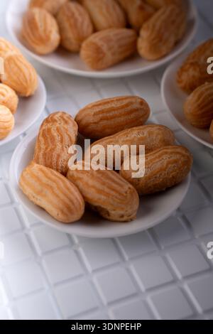 Nahaufnahme goldener Mini-Backwaren, die auf kleinen weißen Tellern über einer weißen Tischplatte angeordnet sind und knusprige geriffelte Muscheln und warme Karamelltöne hervorheben Stockfoto