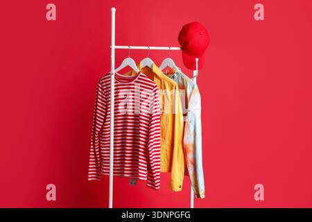 Verschiedene Kleidung und Baseballmütze hängen auf dem Rack vor rotem Hintergrund Stockfoto