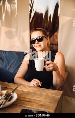 Frau in der Sonnenbrille genießt Kaffee auf der gemütlichen Außenterrasse im warmen Sonnenlicht Stockfoto