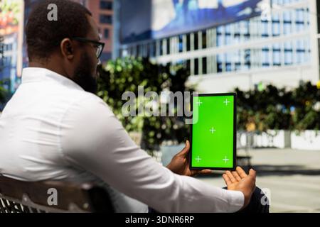 Vertikaler Chroma-Schlüssel mit schwarzem Mann, der entspannt auf einer Bank sitzt und online mit seinem Tablet in der Nähe des Stadtzentrums arbeitet. Straßenstil und selbstbewusste Einstellung im Großstadtleben. Stockfoto
