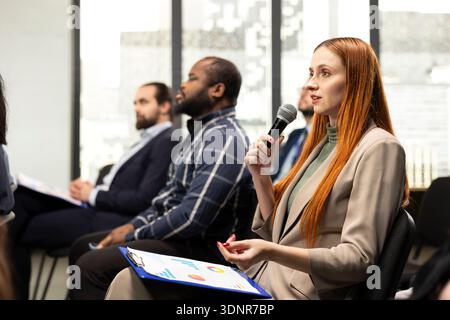 Junge Erwachsene im Publikum, die sich über das Mikrofon an einer Debatte beteiligen und während einer Geschäftsveranstaltung interagieren, während professionelle Personen ihre Meinung in einer Podiumsdiskussion austauschen. Gipfeltreffen des Konvents. Stockfoto