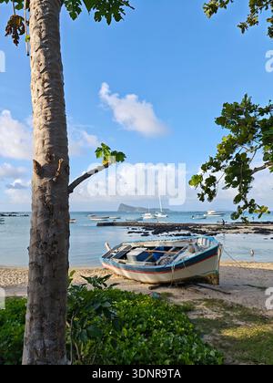 Fischerboot ruht an einem Sandstrand in Mauritius, mit türkisfarbener Lagune, ankerbaren Booten und der Insel Coin de Mire im Hintergrund. Stockfoto