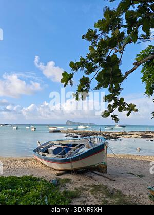 Fischerboot ruht an einem Sandstrand in Mauritius, mit türkisfarbener Lagune, ankerbaren Booten und der Insel Coin de Mire im Hintergrund. Stockfoto