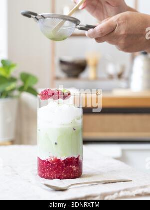 Person, die Grünteepulver über einem erfrischenden Eismatcha Latte mit Himbeeren siebt Stockfoto
