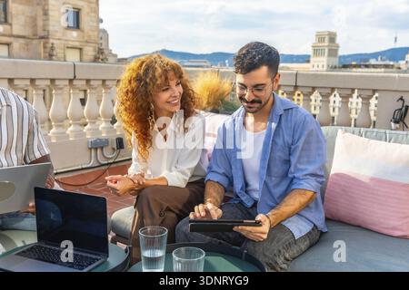 Zwei verschiedene Kollegen arbeiten während eines Business Meetings im Freien auf einem professionellen Coworking Dach zusammen, wobei sie einen Laptop und ein Tablet für Teamarbeit und Teamarbeit verwenden Stockfoto