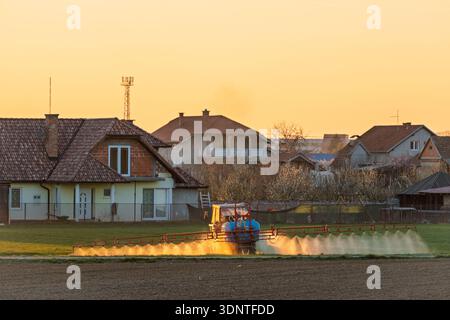 Eine ruhige ländliche Szene bei Sonnenuntergang verfügt über einen Sprinkler, der ein Feld vor Häusern und Gärten bewässert, was das ruhige und praktische Leben auf dem Land einfängt Stockfoto