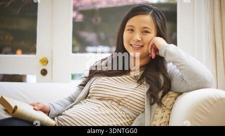 Porträt, Heim- und schwangere Frau auf Sofa, Buch lesen und sich mit Wochenendferien, Hobby und Lächeln entspannen. Glücklich, Literatur und asiatische Person auf der Couch Stockfoto