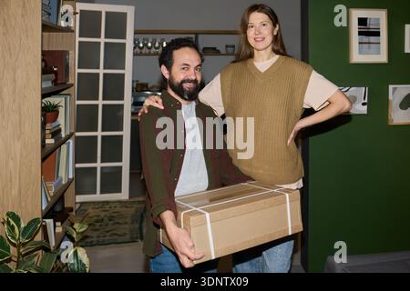 Porträt eines jungen, erwachsenen Kaukasiers, der lächelt, während er einen Karton hält, neben der jungen, erwachsenen Frau des Kaukasiers steht, mit der Hand auf der Hüfte Stockfoto