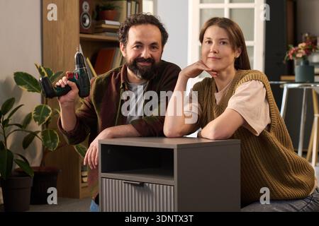 Porträt eines kaukasischen Mannes mittleren Alters mit schnurlosem Bohrgerät neben einer kaukasischen Frau mittleren Alters, die Möbel in einem modernen Wohnraum zusammenbaut Stockfoto