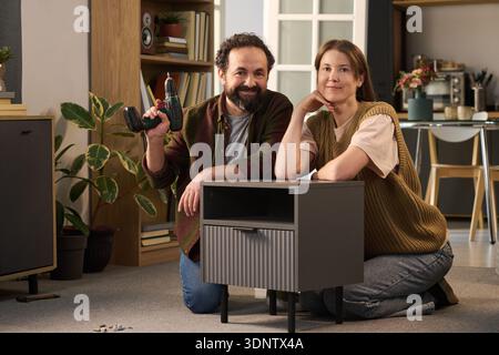 Porträt eines kaukasischen Mannes mittleren Alters, der einen elektrischen Bohrer hält, kniend neben einer kaukasischen Frau mittleren Alters, die einen kleinen Schrank in einem modernen Wohnzimmer zusammenbaut Stockfoto