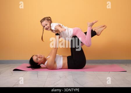 Glückliche Mutter und Tochter üben spielerische Yoga-Posen auf einer rosafarbenen Matte vor orangem Hintergrund, lächeln und verbinden sich miteinander. Stockfoto