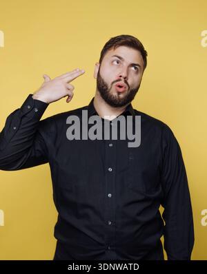 Ein junger Mann mit Bart schlägt selbstbewusst eine lustige Pose und gibt vor, sich mit den Fingern zu erschießen. Er trägt ein schwarzes Hemd und hebt sich von einem ab Stockfoto