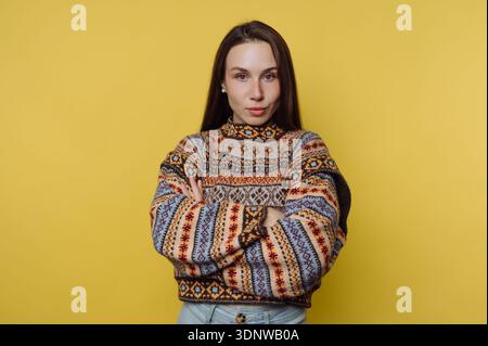 Eine junge Frau steht selbstbewusst mit überkreuzten Armen und zeigt einen kuscheligen, farbenfrohen Pullover. Ihr Ausdruck spiegelt Selbstbewusstsein wider. Das helle Gelb Stockfoto