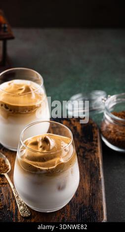 Cremiger Dalgona-Kaffee, serviert in eleganten Gläsern auf einem rustikalen Holzbrett mit Kaffeesatz Stockfoto