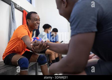Afroamerikanische Männer mit Knieschützern, die Hände auf einer Bank in der Umkleidekabine schütteln und Volleyball halten. Spind, Athlet, Teamkollege, Ball, Teamwork, offen, li Stockfoto