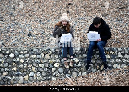 Brighton, Vereinigtes Königreich - 13. Januar 2018: Zwei Freunde essen Fish and Chips zum Mitnehmen, während sie an einem kalten Wintertag auf einer Kieselsteinmauer sitzen Stockfoto