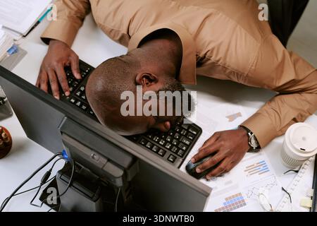 Junger schwarzer Mann, der während des Arbeitstages auf dem Schreibtisch schläft Stockfoto