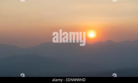 Ein trüber Sonnenuntergang über den mehrschichtigen Bergkämmen während der brennenden Jahreszeit in Pai, Nord-Thailand, mit warmem Sonnenlicht, das durch den rauchgefüllten Himmel strahlt. Stockfoto