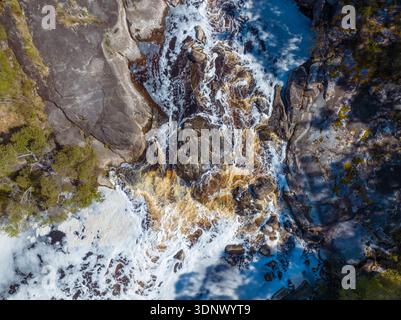 Aus der Luft blitzschnell bewegendes Flusswasser, das über dunkle Felsformationen schäumt und wirbelnde Muster in einer bewaldeten Schlucht erzeugt. Stockfoto