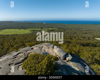 Aus der Vogelperspektive ist ein einsamer Wanderer auf einem Granitgipfel mit Blick auf weitläufige Wälder und ferne Küste zu sehen. Stockfoto