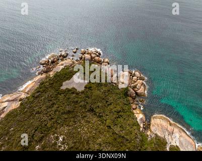 Ein Blick aus der Vogelperspektive auf eine zerklüftete Landzunge mit dichter Vegetation und abgerundeten Granitblöcken, die auf klares türkisfarbenes Wasser treffen. Stockfoto