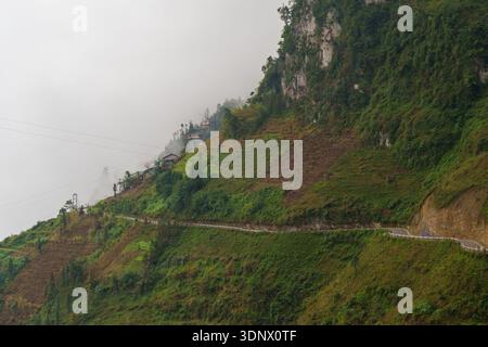 Eine schmale Bergstraße führt entlang steiler grüner Klippen auf einem nebeligen Hügel entlang der Ha Giang-Schleife in der Nähe von Dong Van. Stockfoto