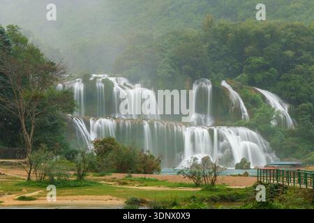 Der Ban Gioc Wasserfall fällt über mehrere Ebenen in üppige Wälder, mit einer Fußgängerbrücke, die den Fluss überquert. Nebel steigt von t auf Stockfoto