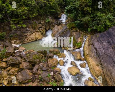 Kleiner Wasserfall, der durch einen felsigen Bach fließt, umgeben von dichtem grünen Wald. Das Wasser kaskadiert über Felsbrocken in ein flaches Becken. Stockfoto