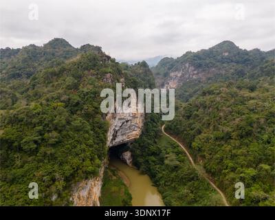 Ein Fluss fließt durch die Puong Cave unter hoch aufragenden Kalksteinklippen, umgeben von bewaldeten Karsthügeln. Stockfoto