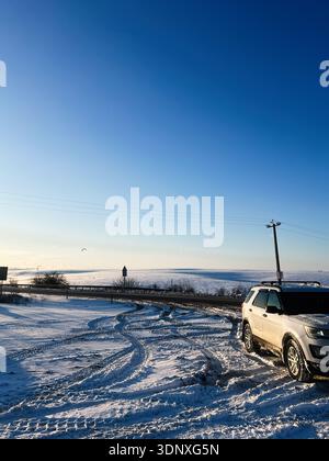 Weißer SUV parkte auf einer schneebedeckten Straße neben einem weiten, klaren blauen Winterhimmel und einer offenen Landschaft. Stockfoto