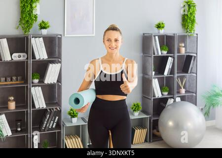 Lächelnde Frau, die die Daumen hoch hält, während sie eine Yogamatte in einem hellen Fitnessraum hält Stockfoto