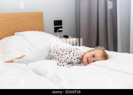 Ein verspieltes Kleinkind mit blonden Haaren liegt fröhlich auf einem gemütlichen Bett, trägt ein Polka-Dot-Top und eine weiße Hose, umgeben von weicher Bettwäsche in einem hellen R Stockfoto
