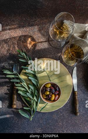 Blick von oben auf einen Tisch für eine Party mit griechischen Oliven in einer Schüssel auf einem grünen Teller, begleitet von Bernsteinwein in Gläsern, vor einer rustikalen Braue Stockfoto