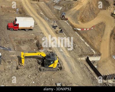Luftaufnahme einer Baustelle mit einem gelben Planierschild, der aktiv Erde gräbt und bewegt, neben einem roten Lkw, der Maschinen und Sitzen zeigt Stockfoto