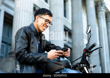 Ein hispanischer Mann in einer schwarzen Lederjacke sitzt auf seinem Motorrad und lächelt, während er sein Smartphone benutzt. Die urbane Kulisse bietet eine beeindruckende Architektur Stockfoto