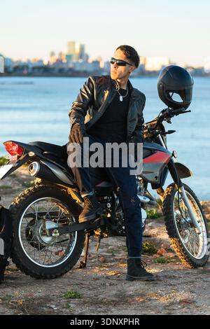 Ein hispanischer Mann, gekleidet in Lederjacke und Sonnenbrille, sitzt selbstbewusst auf einem Motorrad am Wasser. Die Skyline der Stadt und das ruhige Wasser bilden die Grundlage Stockfoto