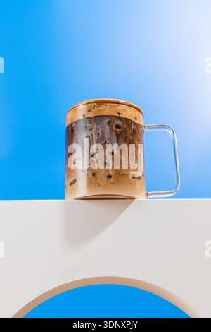 Eine Tasse aus klarem Glas, gefüllt mit Eiskaffee, mit wirbelnden Milchmustern, ruht auf einem modernen weißen Tisch, der vor einer leuchtenden, sonnigen blauen Ske gefangen gehalten wird Stockfoto