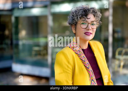 Selbstbewusste Geschäftsfrau mit lockigem Haar, Brille und einer leuchtend gelben Jacke mit farbenfrohen Akzenten, die im Freien mit einem nachdenklichen Auftritt steht Stockfoto