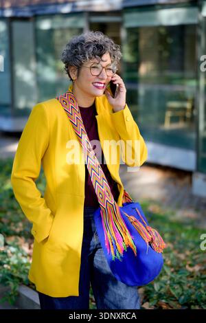 Glückliche Geschäftsfrau, die draußen steht, am Telefon spricht und einen sonnigen Tag genießt. Sie trägt eine leuchtend gelbe Jacke, bunte Accessoires und ich Stockfoto