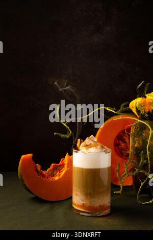 Ein warmer Latte mit Kürbisgewürzen und schaumiger Creme wird kunstvoll neben leuchtenden orangen Kürbisscheiben auf dunklem Hintergrund platziert, was an einen gemütlichen Herbst A erinnert Stockfoto