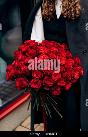 Eine Frau mit lockigem Haar in einem stilvollen Outfit hält einen großen, leuchtenden Blumenstrauß roter Rosen. Das elegante Arrangement bildet einen markanten Kontrast zu ihr Stockfoto