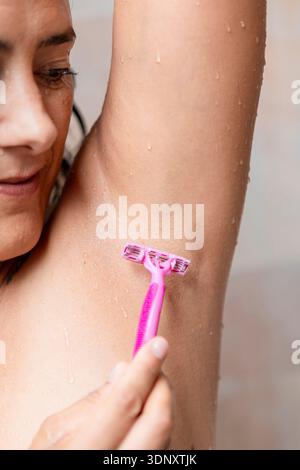 Ein Nahaufnahme-Bild einer Frau, die ihre Achselhöhle sanft mit einem rosa Rasierer unter der Dusche rasiert. Wassertropfen sind sichtbar, was auf einen frischen und sauberen Routin hinweist Stockfoto