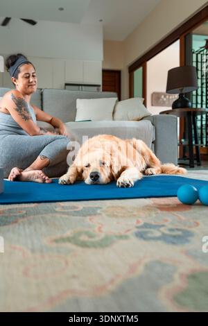 Eine Frau in sportlicher Kleidung sitzt zu Hause mit ihrem Golden Retriever auf einer Yogamatte in entspannter Pose. Der Hund ruht sich ruhig aus und sorgt für einen friedlichen Ambi Stockfoto