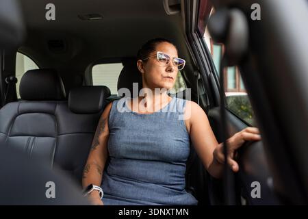 Eine Geschäftsfrau im Kleid sitzt auf dem Rücksitz eines Autos und blickt nachdenklich aus dem Fenster. Das natürliche Licht unterstreicht ihren fokussierten Ausdruck und Stockfoto