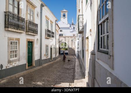 Eine Frau geht mit ihrem Hund durch eine charmante Kopfsteinpflasterstraße in Faro, Algarve, Portugal. Die historische Architektur und die hellen Gebäude bilden ein Bild Stockfoto