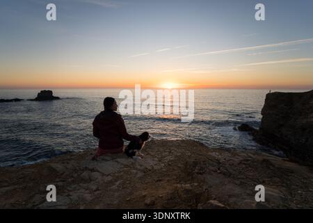 Eine Frau sitzt neben ihrem Hund auf einer Klippe in Zambujeira do Mar, Portugal, und bestaunt einen atemberaubenden Sonnenuntergang über dem Atlantischen Ozean. Die Küste der Algarve Stockfoto
