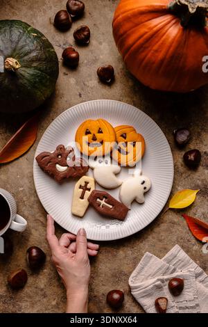 Blick von oben auf beschnittene, nicht erkennbare Menschenhände mit dekorierten Halloween-Keksen auf einem weißen Teller, umgeben von Kürbissen, Kastanien und farbenfrohen Lev Stockfoto