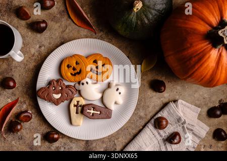 Blick von oben auf Halloween-Kekse in Form von Kürbissen, Geistern, Fledermäusen und Gräbern auf einer weißen Platte. Umgeben von Kastanien, Kürbissen und einer Tasse Co Stockfoto